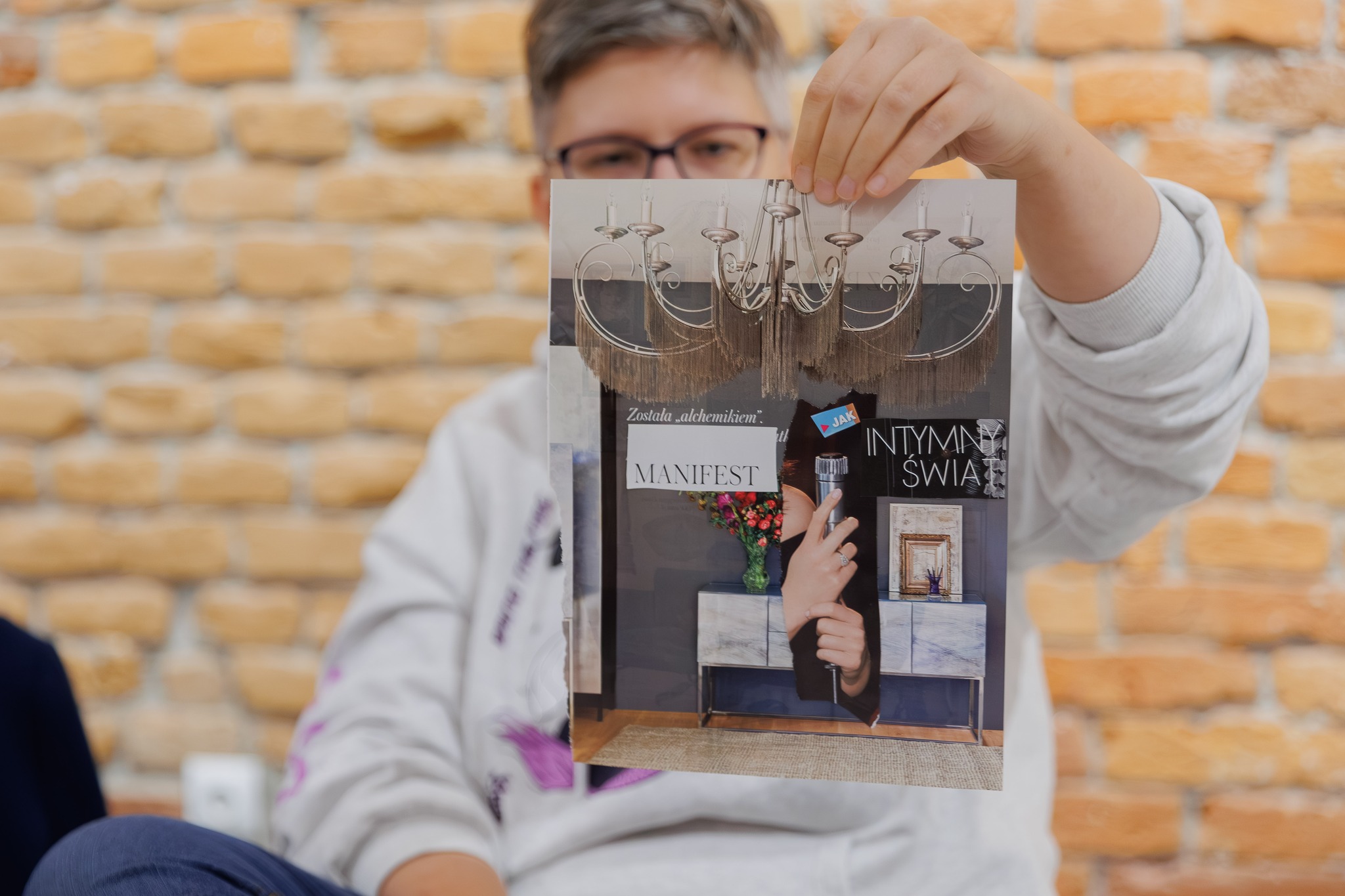 Kobieta w okularach prezentuje kolaż intuicyjny z żyrandolem, kwiatami, mikrofonem i napisem „MANIFEST”.