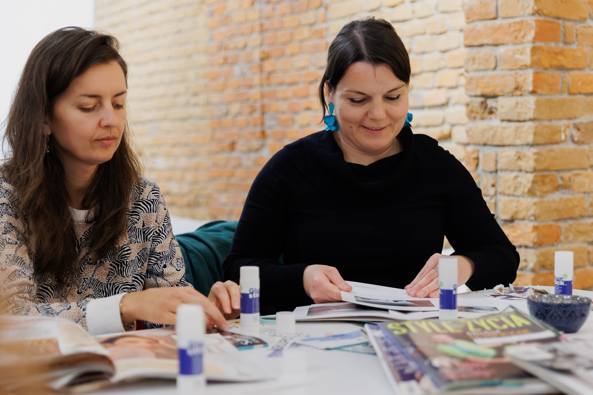 Dwie kobiety przyklejają wycinki na kolaż intuicyjny przy stole z czasopismami, papierowymi fragmentami i klejem w sztyfcie.